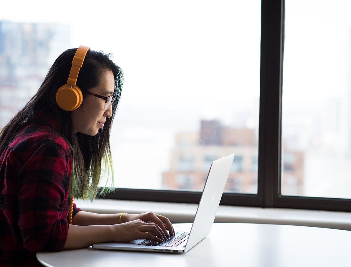 Image of woman wearing yellow headphones working on laptop in front of office building window A woman wearing headphones is typing on a laptop computer.