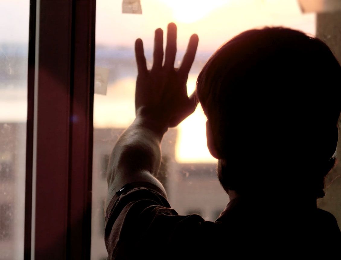 A person is looking out of a window with their hand on the glass.