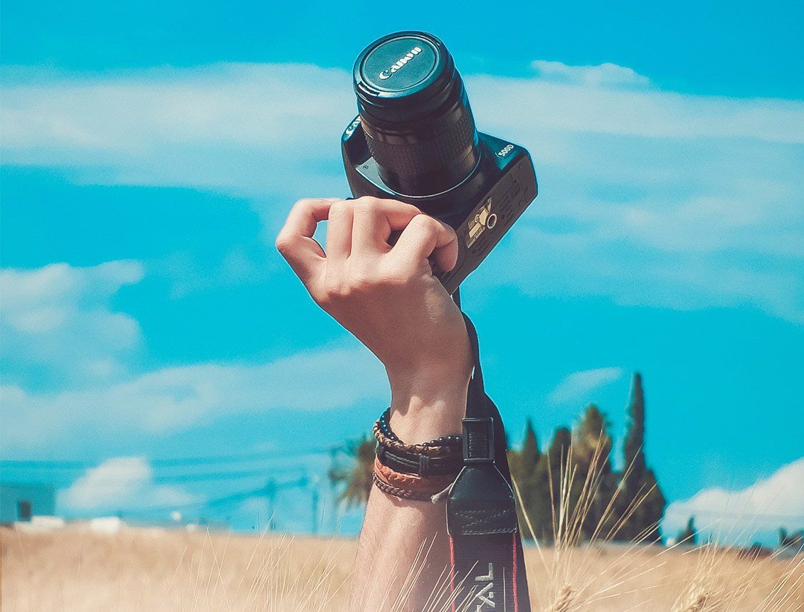 Close up hand holding up DSLR video camera in field with blue sky behind A person is holding a camera in their hand in a field.