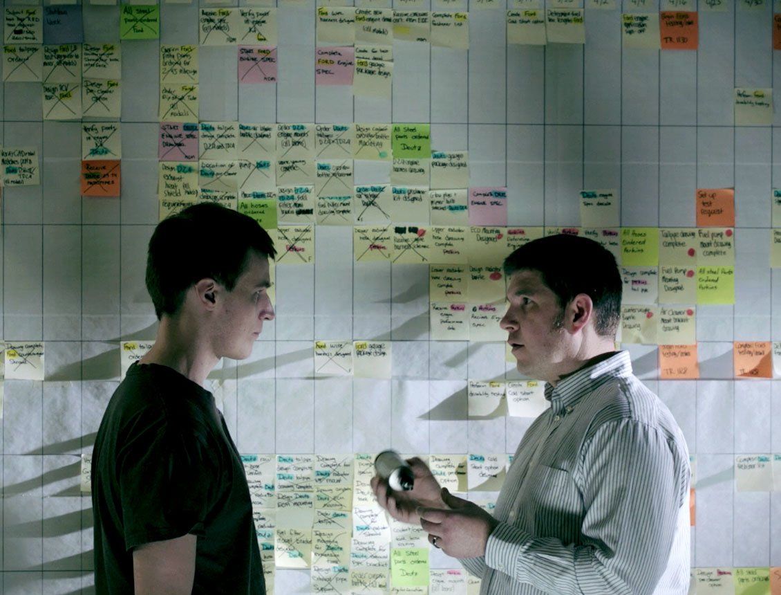 Two men are standing in front of a wall with sticky notes on it