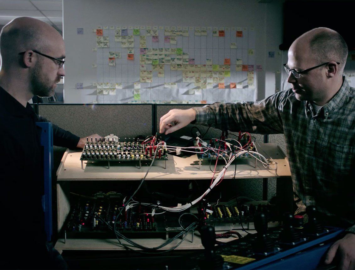 Two men in glasses working on complex electronics with wires and a control panel.