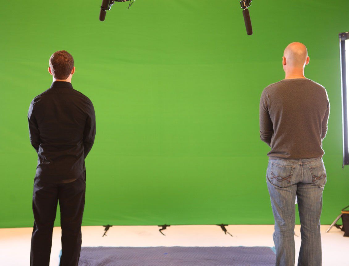 Two people standing in front of a green screen; microphones overhead.