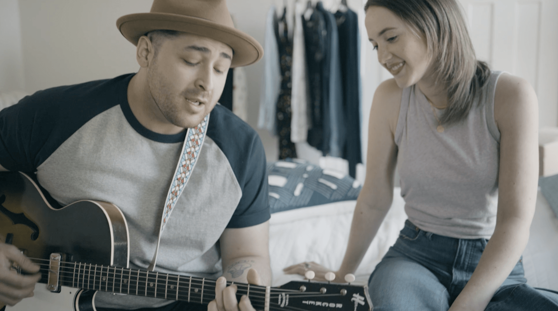 Man plays guitar in bedroom as girlfriend listens and looks at him playing A man is playing a guitar for a woman who is sitting on a bed.