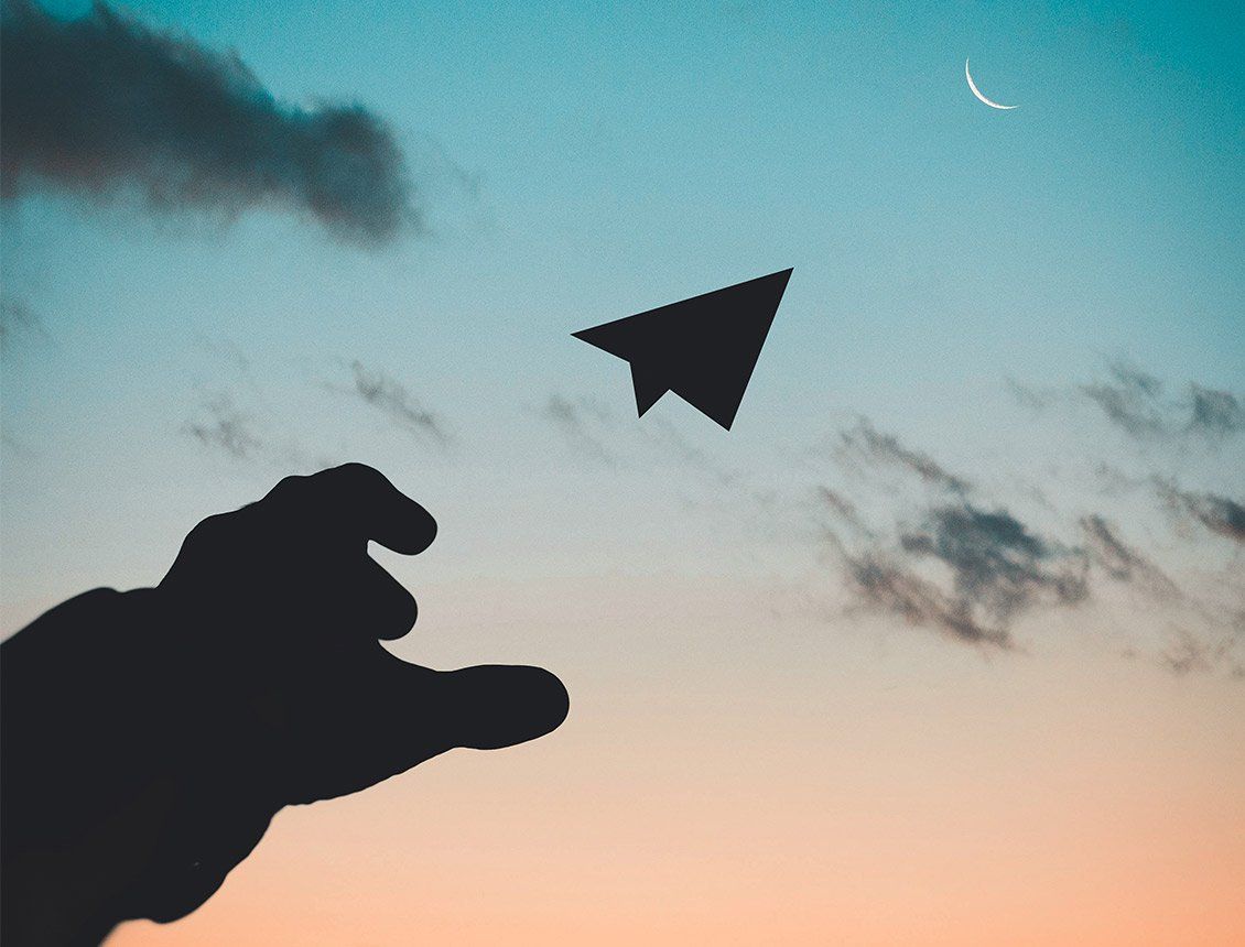 Silhouette of a hand throwing a paper airplane into a dusk sky with a crescent moon and clouds.