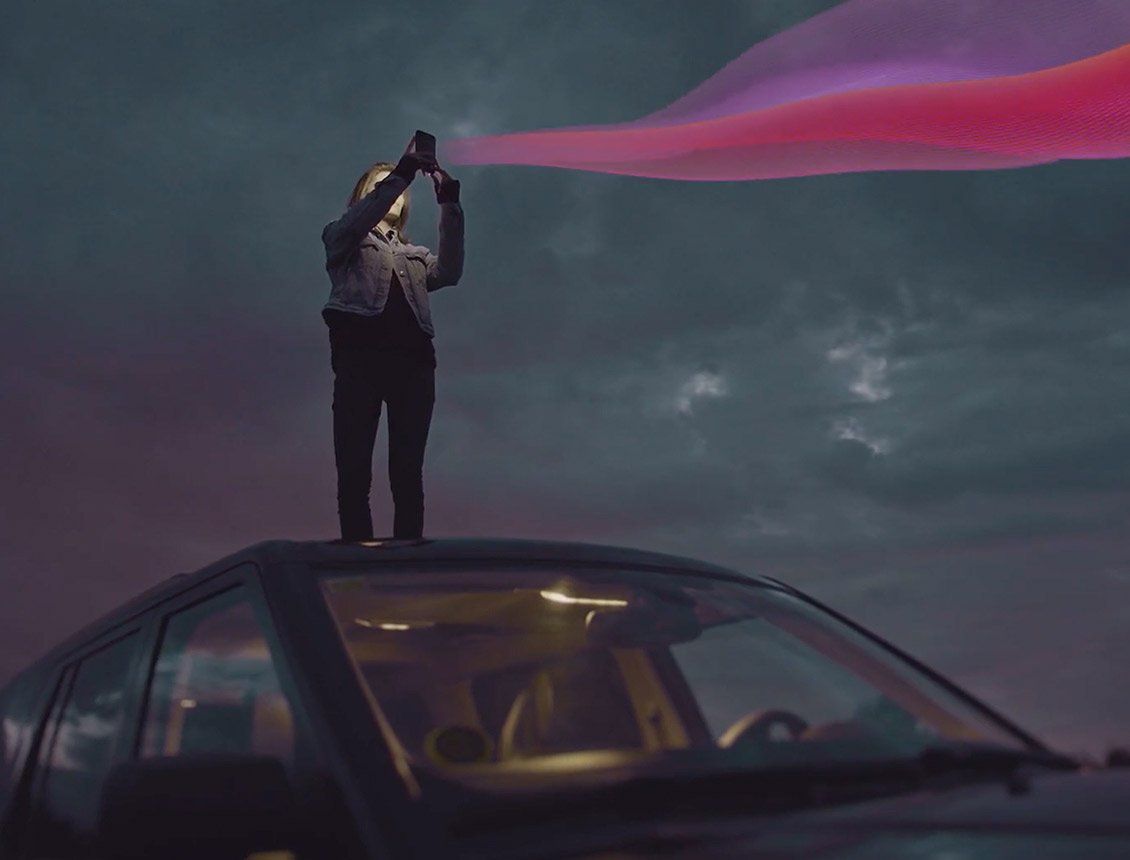 Image of woman at night on top of car looking at mobile device with pink graphic ribbon coming out of phone Person standing on a car roof, holding a device emitting a pink/purple beam into a stormy sky.
