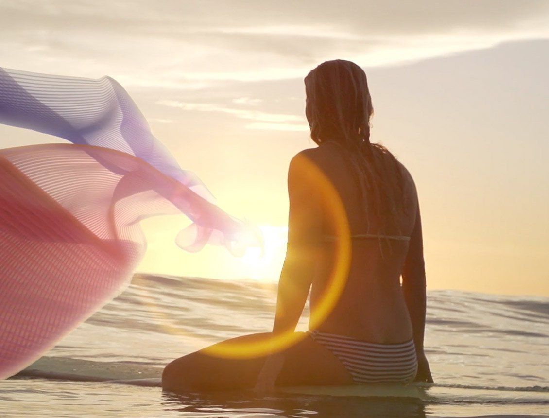 Image of women on surfboard on the ocean with a pink ribbon graphic beside her at sunset Woman on surfboard at sunset, looking toward the sun; fabric blowing in the wind.