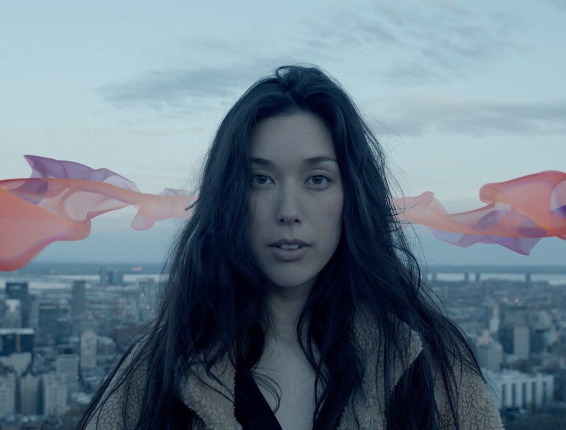 Image of woman's portrait looking into camera lens with pink graphic ribbon behind her in front of a large city Woman with dark hair looking at camera, cityscape backdrop; flowing orange fabric.