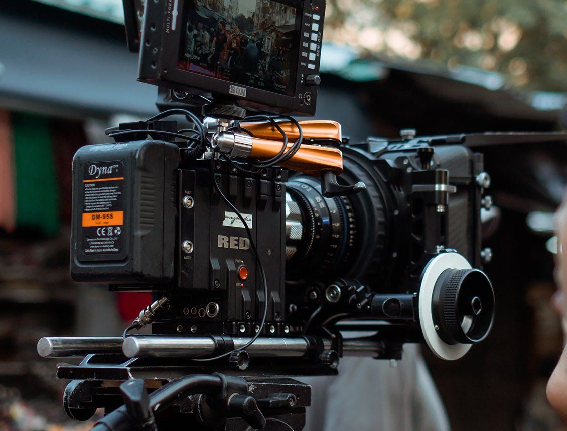 Black professional video camera on a tripod, with attached monitor and battery. Outdoors, partially blurred background.