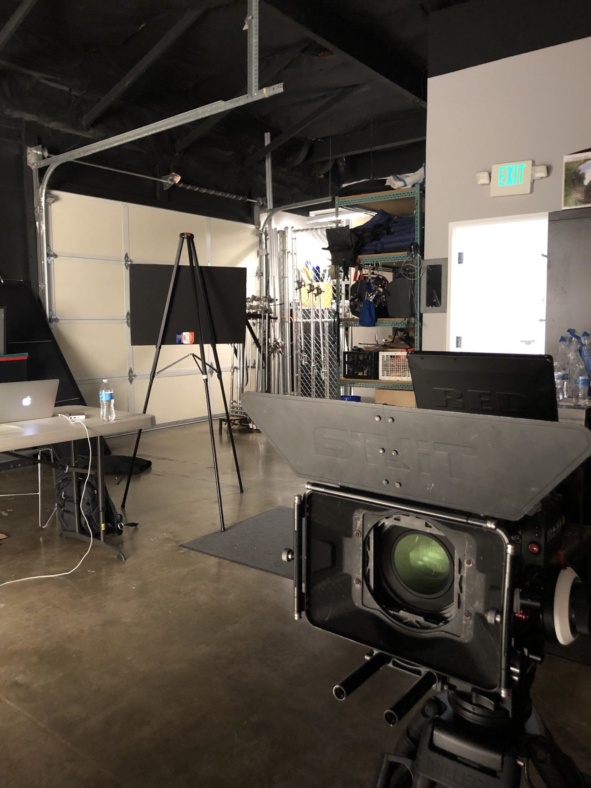 Film set with camera in foreground, garage door, equipment, and crew workspace.