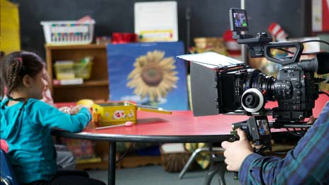 Spin Creative behind the scenes photo Treehouse brand video tv commercial shoot girl sitting in classroom A man is holding a camera in front of a little girl sitting at a table.