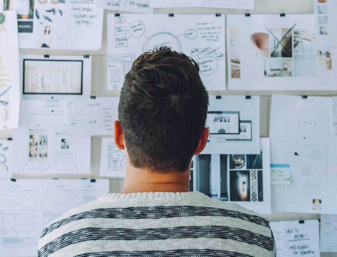 Person with dark hair and striped sweater looking at a wall covered in papers, diagrams, and sketches.