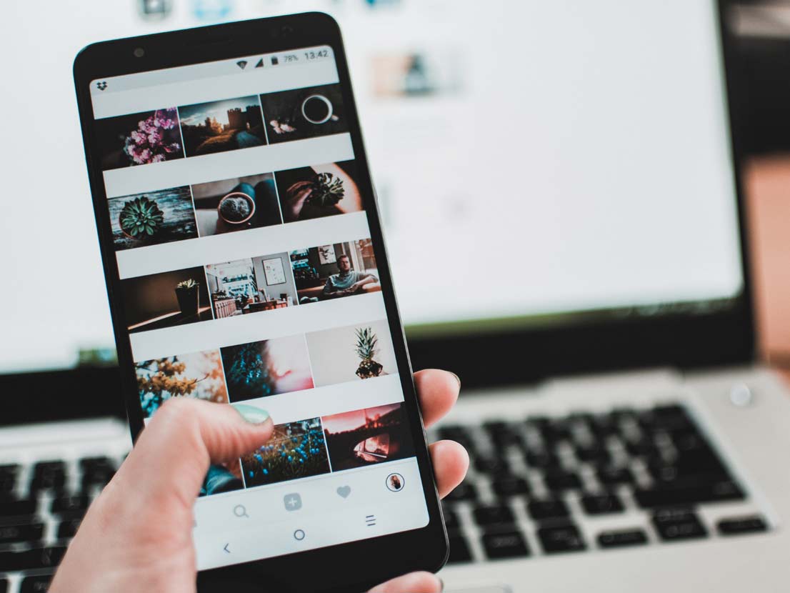 Hand holding a black smartphone displaying a social media feed, laptop in the blurred background.