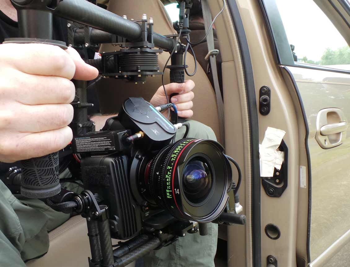 Camera rig inside a car, held by a person. Black camera with a lens. Beige car interior.