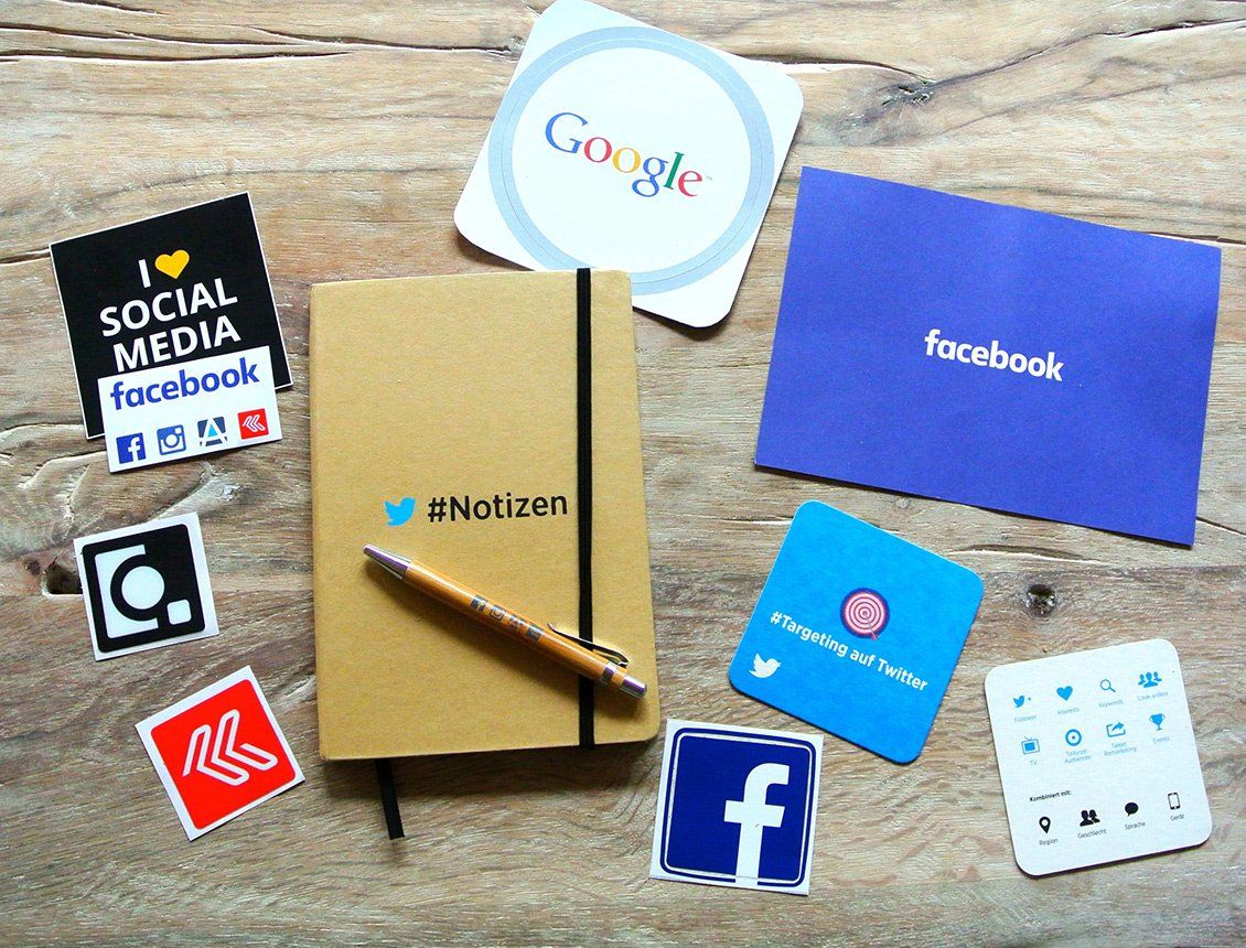 Social media logos including Facebook, Google, Instagram, and Twitter surround a notebook and pen on a wooden surface.