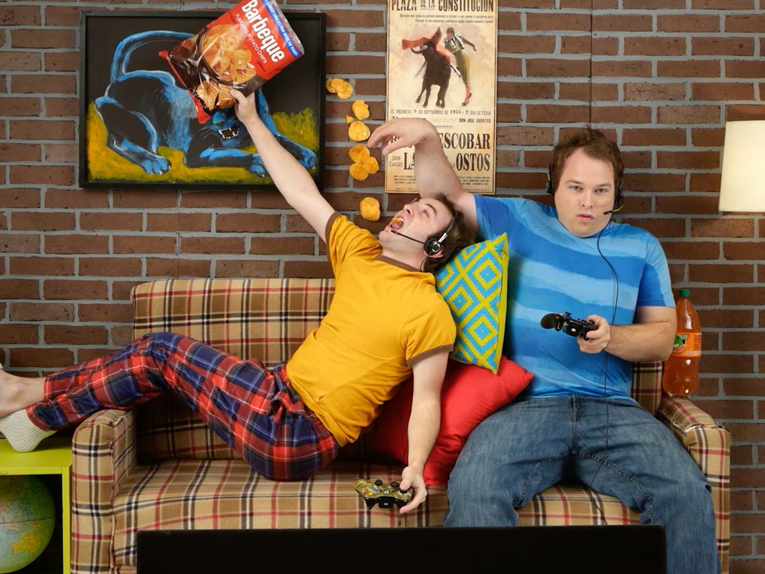 Two men are sitting on a couch eating chips and playing video games