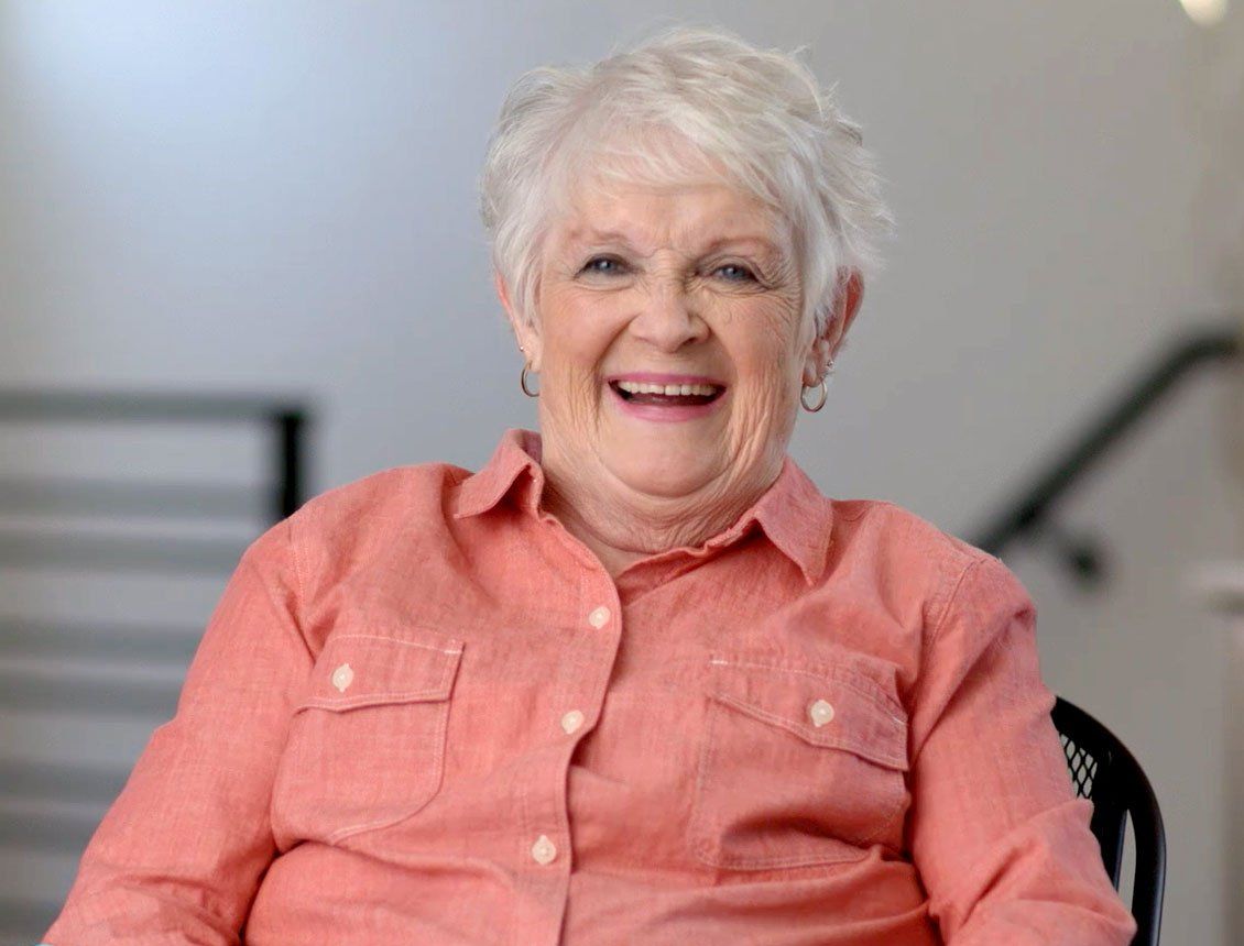 An elderly woman is sitting in a chair and smiling for the camera.