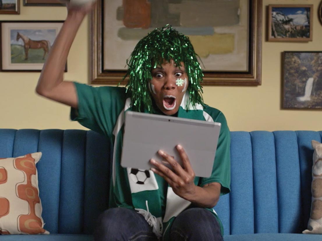 Person cheering while watching a match on a tablet, wearing a wig, scarf, and face paint, seated on a blue couch.