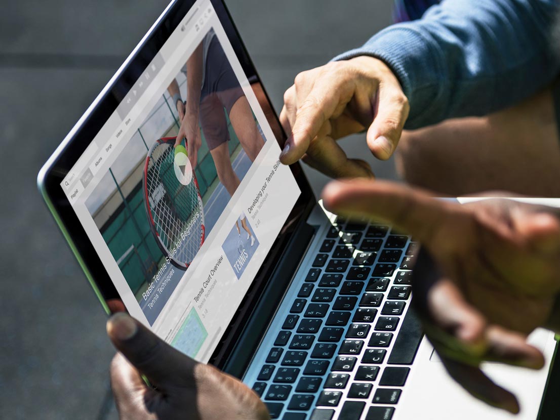 Two people are looking at a laptop screen with a tennis player on it