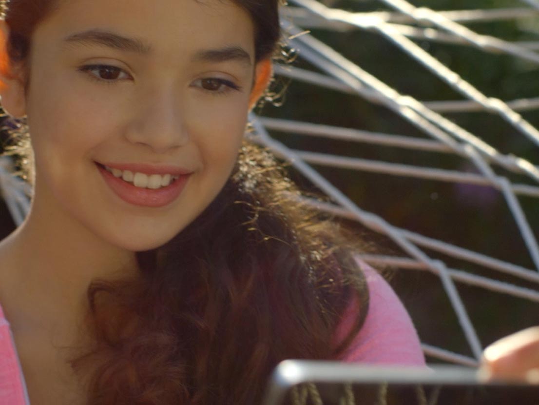 Girl smiling, looking at a tablet, outdoors. She wears a pink shirt. Sunlight.