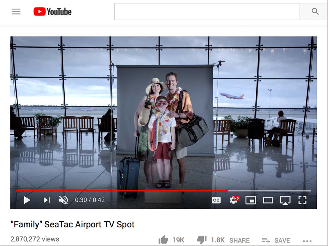 Family posing at airport, wearing vacation attire. Airport background with windows and a plane in the distance.