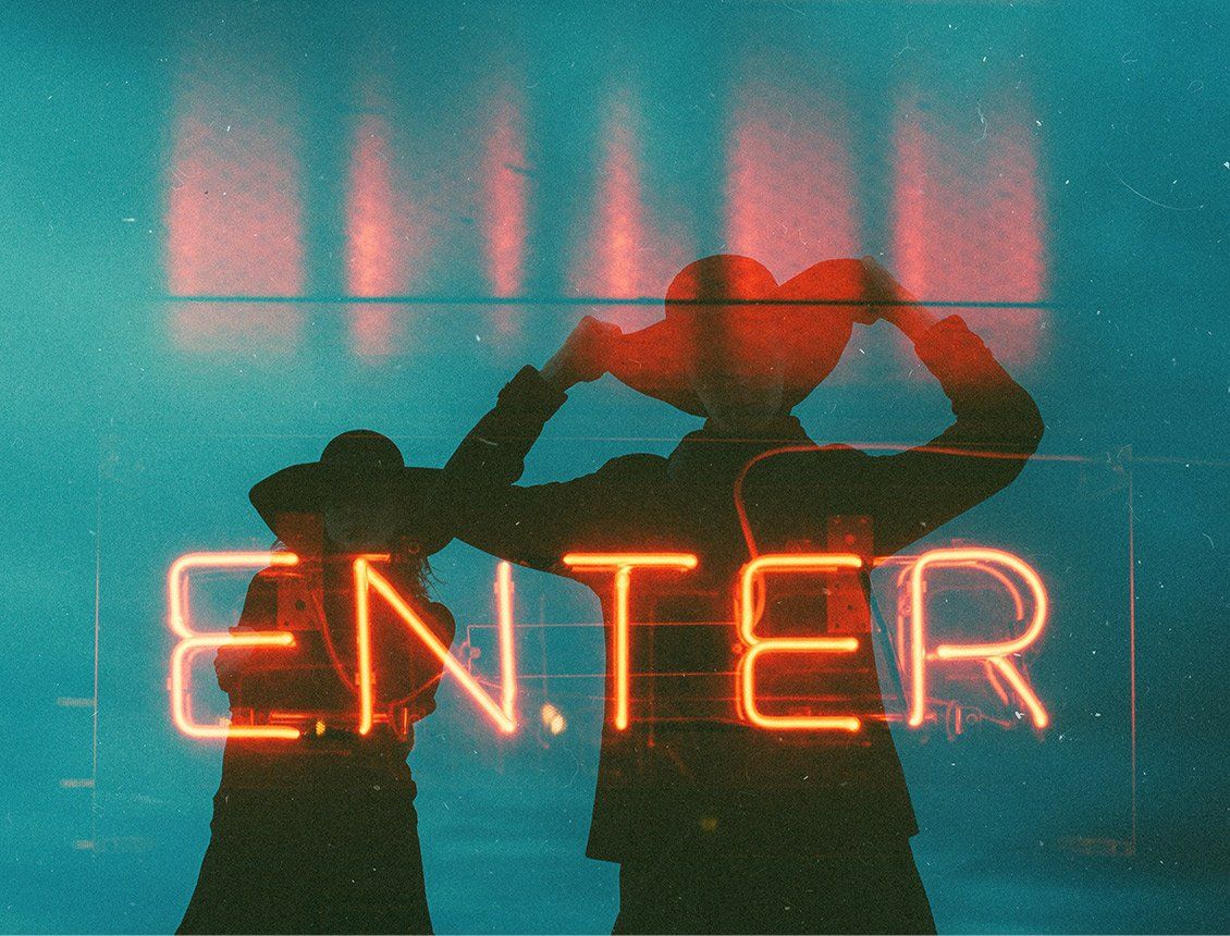 Sillouhette image of two people wearing cowboy hats behind neon Two people are standing in front of a neon sign that says enter