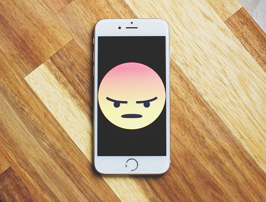 Top down close up of phone sitting on wooden table with sad face on screen White smartphone displaying an angry emoji on a wooden surface.