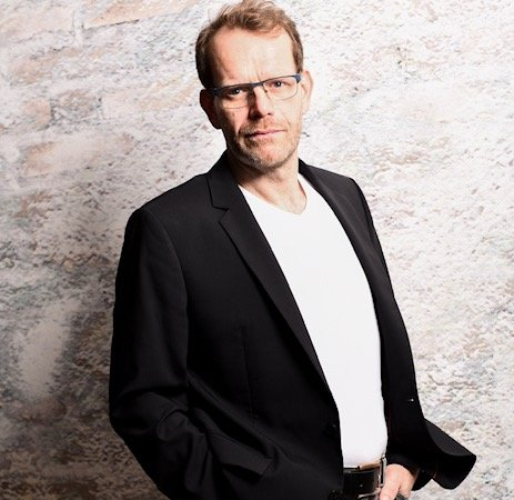 Ein Mann mit Brille und schwarzem Anzug steht vor einer Backsteinmauer