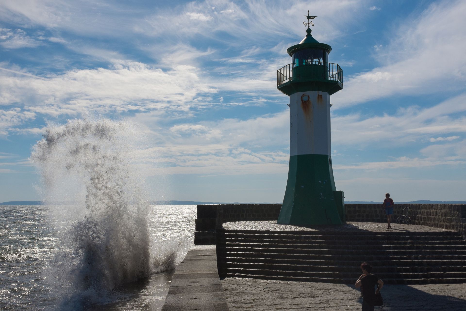 Ein grün-weißer Leuchtturm auf einem Pier, gegen den die Wellen schlagen