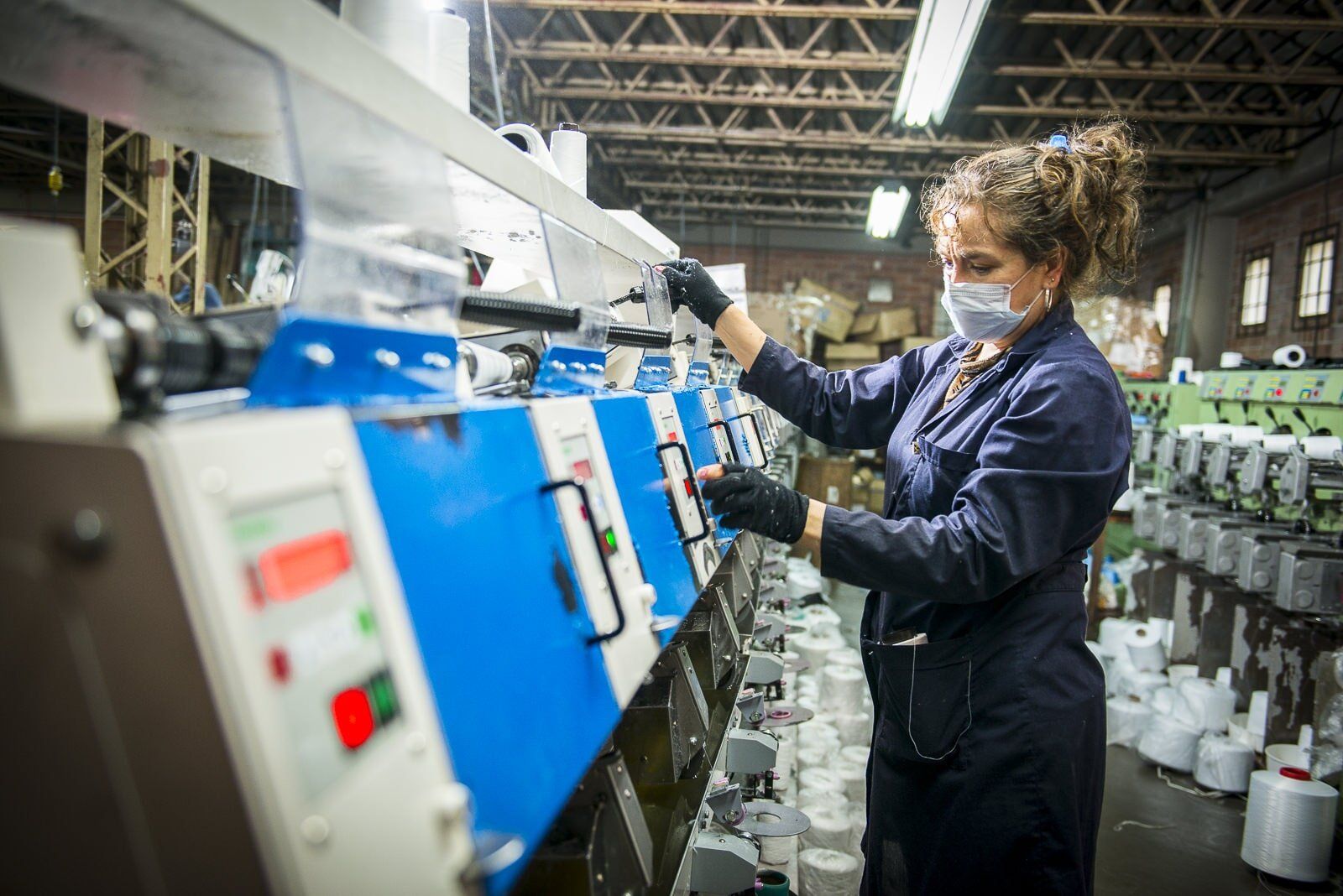 Worker in dark blue coveralls inspecting blue machinery in a factory line.