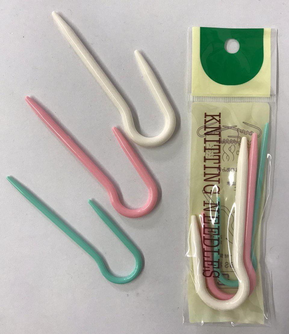 Three U-shaped knitting stitch holders in white, pink, and green beside packaged holders on a card.