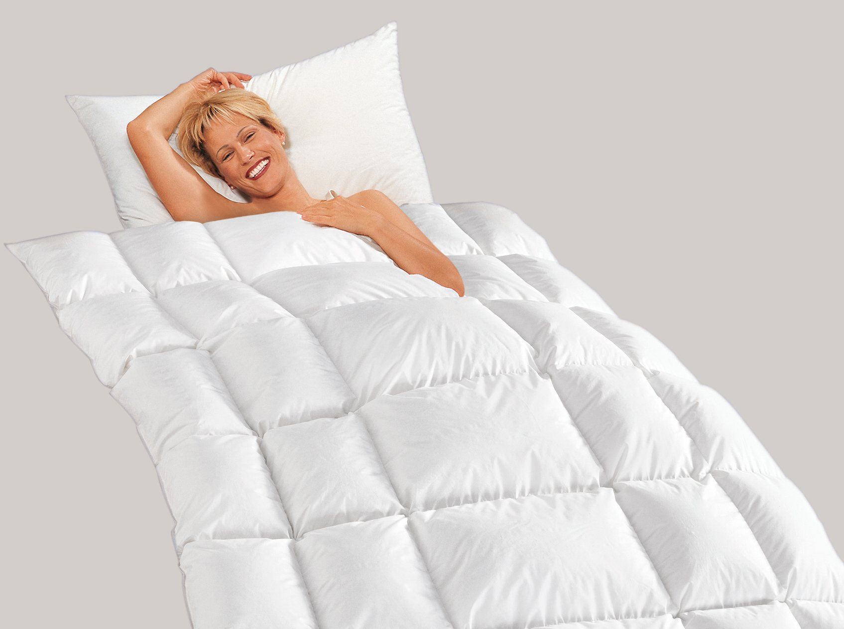 Lächelnde Frau im Bett unter einer weißen, gesteppten Bettdecke und einem Kissen. Heller Hintergrund.