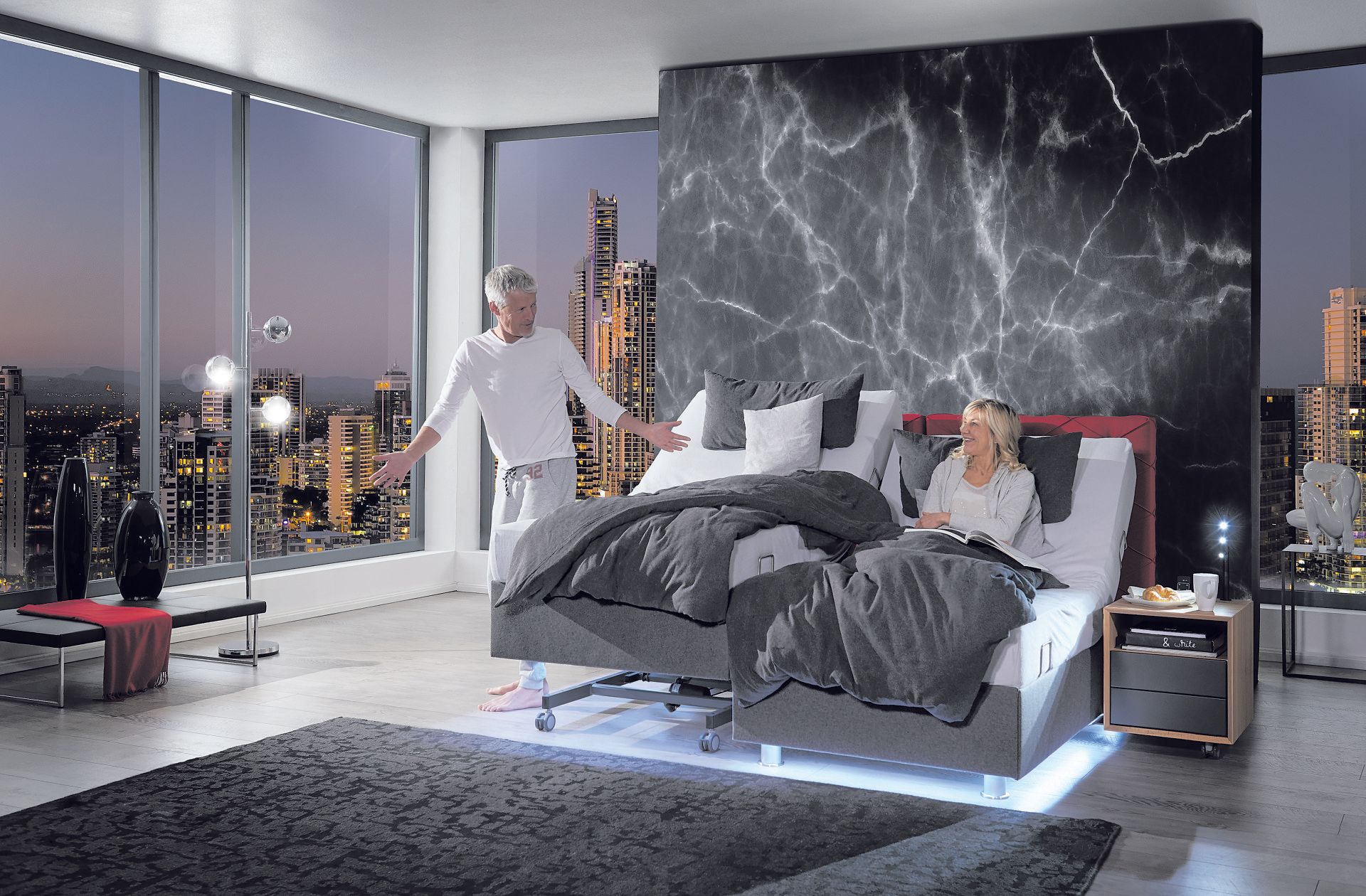 Mann und Frau in einem modernen Schlafzimmer mit verstellbarem Bett, beleuchtet von den Lichtern der Stadt.