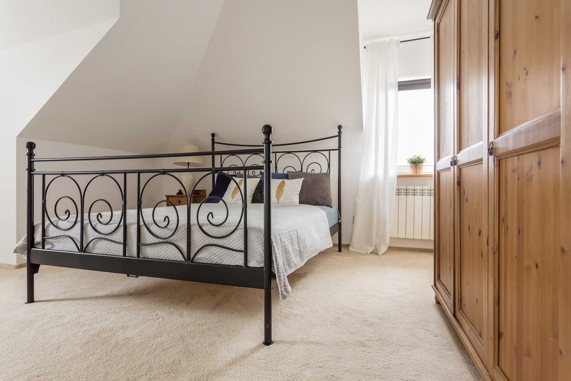 Schlafzimmer mit schmiedeeisernem Bett, hellem Teppich und Holzschrank. Ein Fenster mit Vorhängen ist sichtbar.