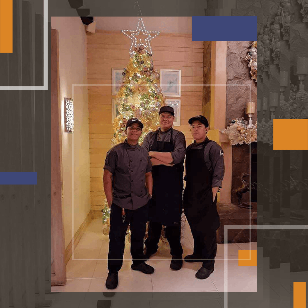 Three people standing indoors beside a lit Christmas tree, smiling in a hallway.