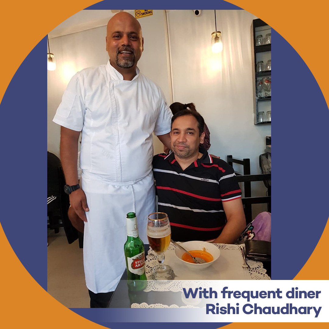 Two men at a restaurant table with drinks and soup; one stands in white chef attire, with text “With frequent diner Rishi Choudhary”
