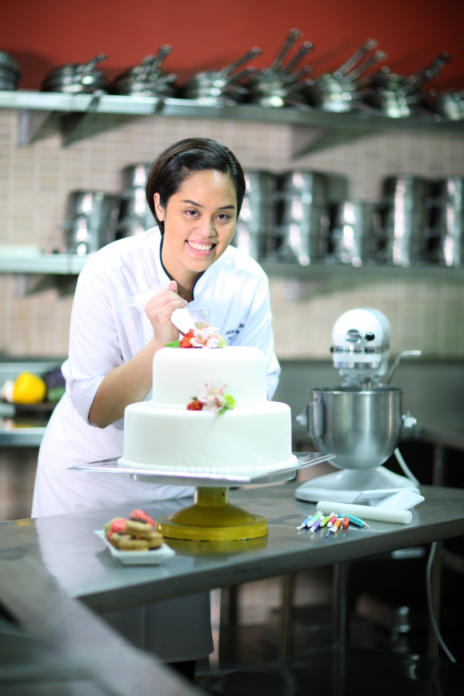 Chef decorating a white cake in a kitchen with a stand mixer and metal shelves