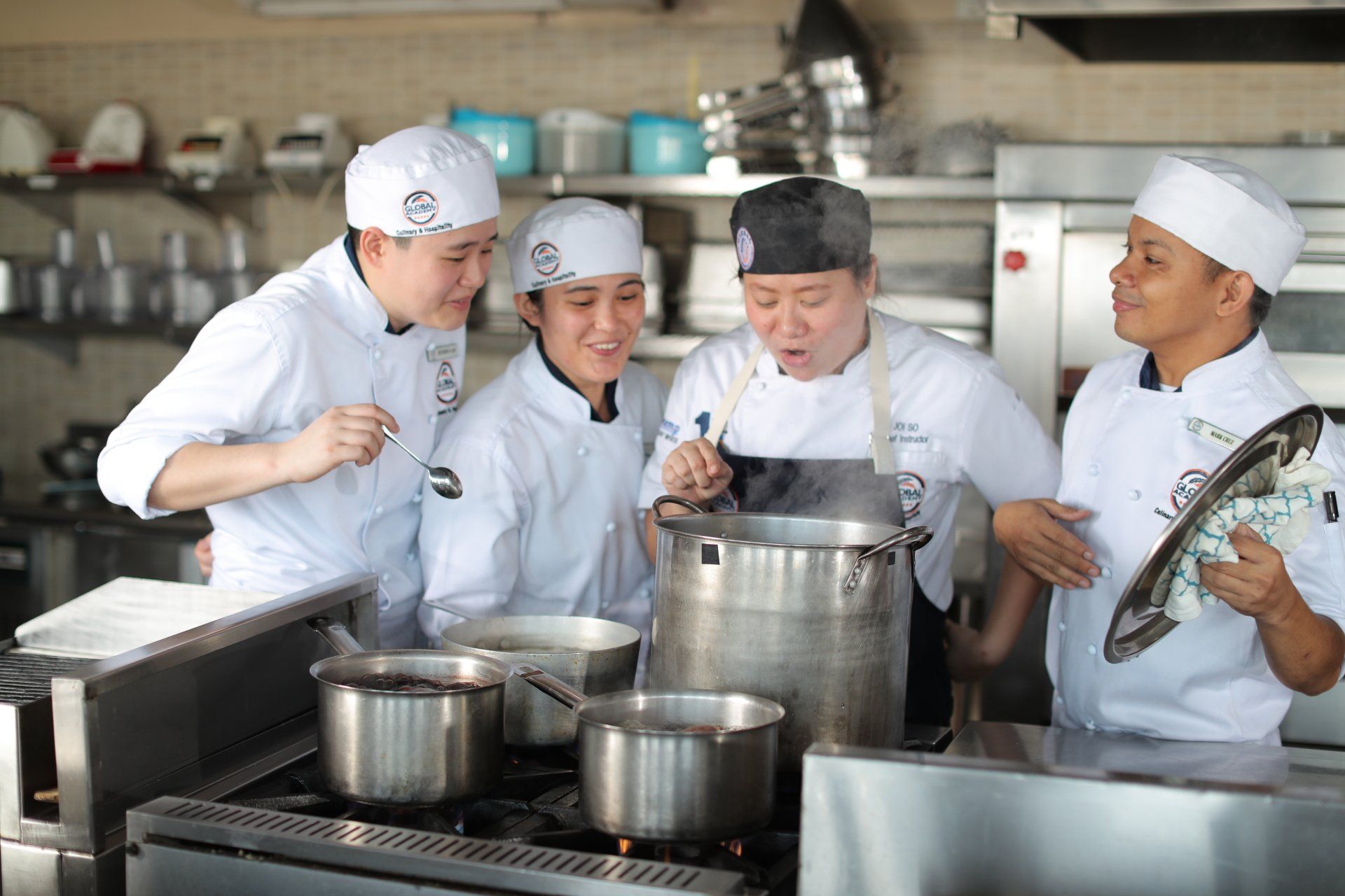 Chefs in white uniforms cooking together in a commercial kitchen, stirring a large pot and smiling
