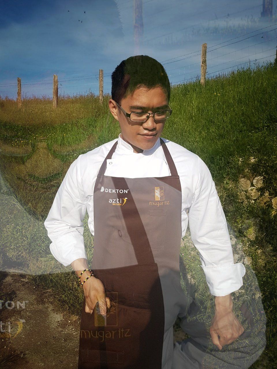 Person in a white shirt and brown apron standing outdoors in a grassy field under a blue sky.