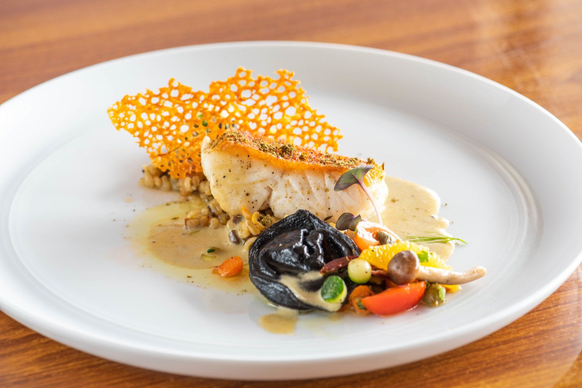 Gourmet plated fish with orange lace crisp, black olive garnish, and colorful vegetables on a white plate