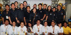 Group photo of people in dark and white shirts posing indoors with medals and balloons in the background