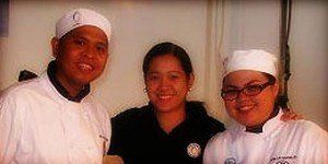 Three people in chef uniforms smiling indoors, with the center person in a dark shirt.