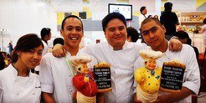 Four smiling people in chef uniforms holding stuffed toys and signs at a bright indoor event