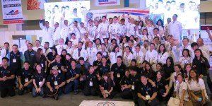 Large group posing for a formal team photo on a stage with a white backdrop and banners