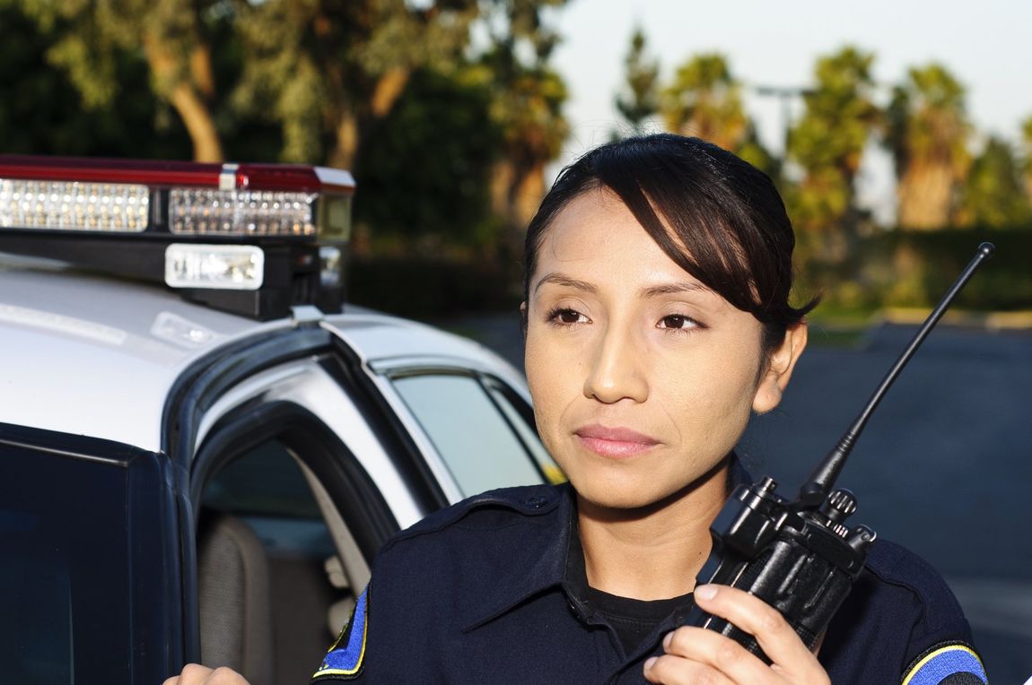 Police officer holding a walkie-talkie beside a patrol car with emergency lights on
