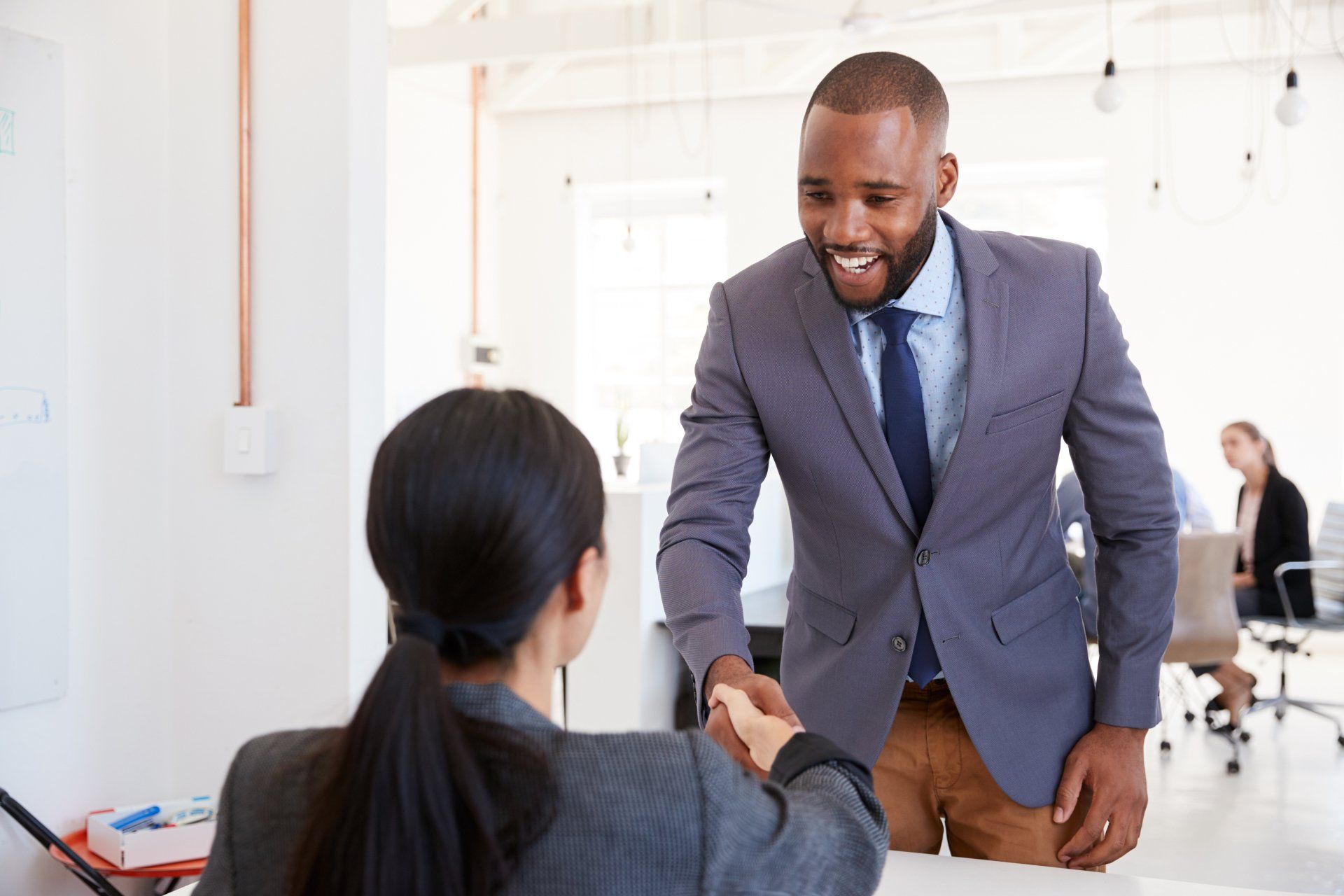 Handshake between two business professionals in a bright office, one standing in a blue suit and smiling.