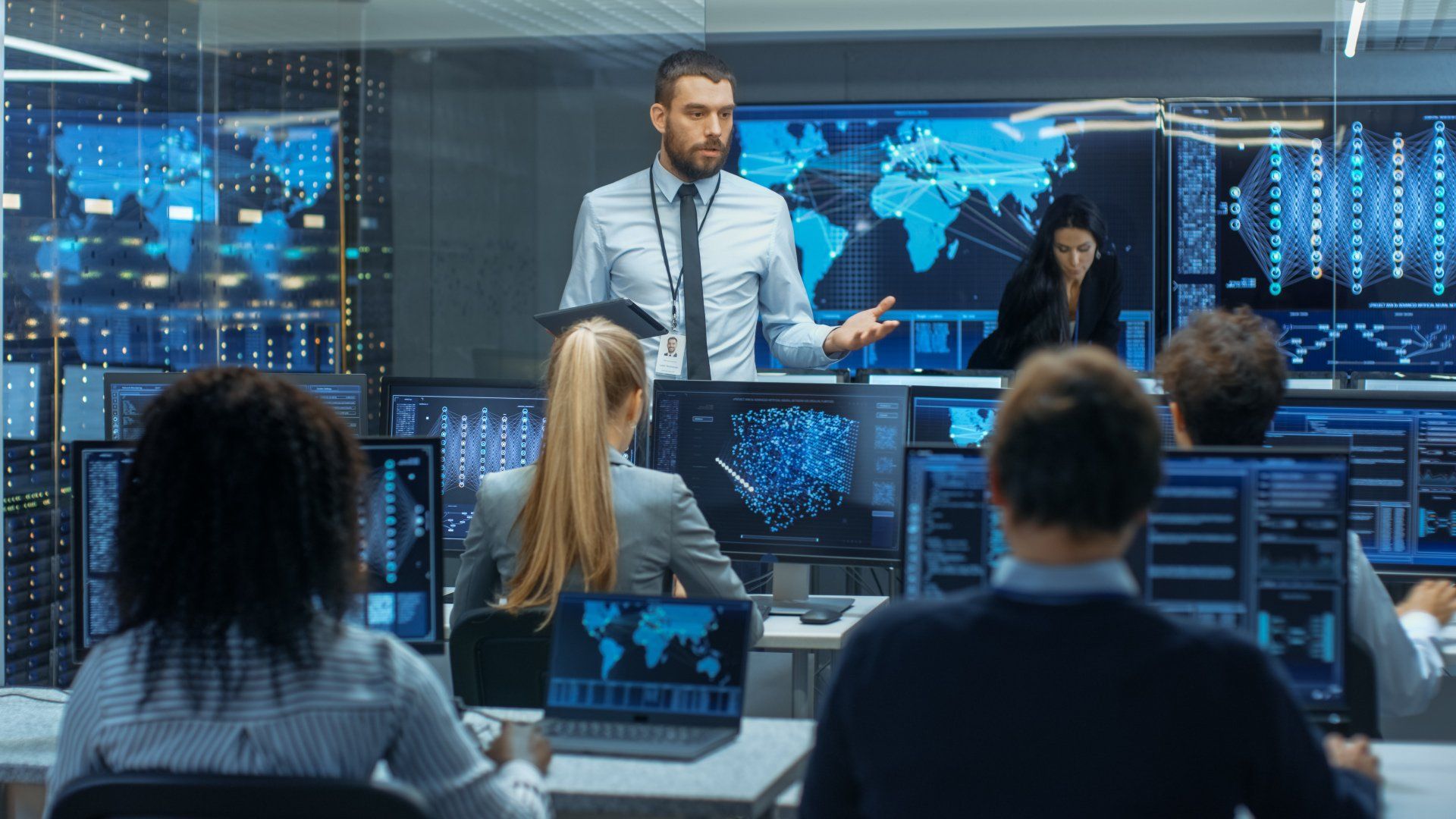 Team in a blue-lit control room analyzing data on multiple monitors while a presenter gestures at a wall display