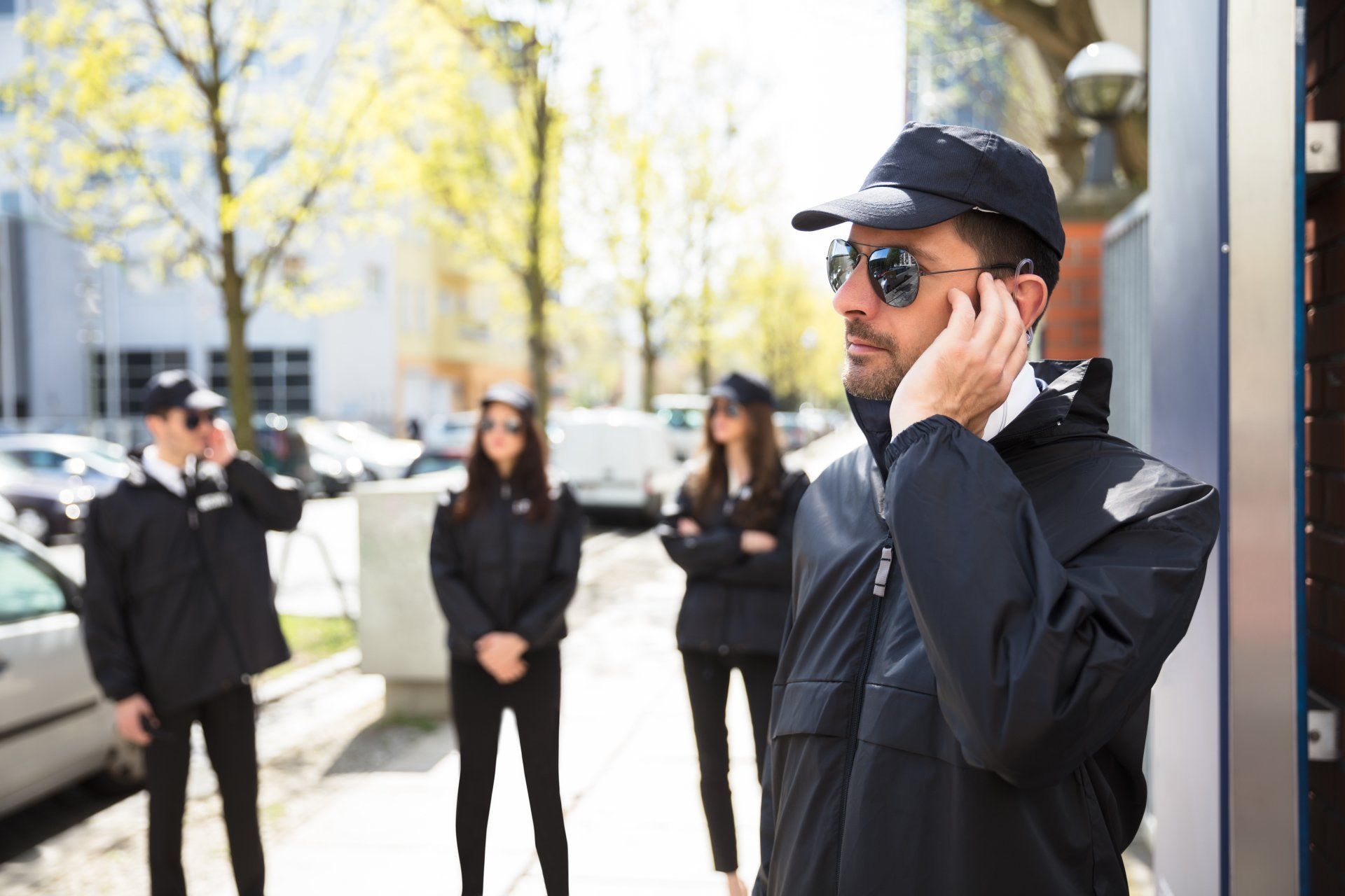 Man in black jacket and cap using a phone outside a building, with three blurred people in the background.