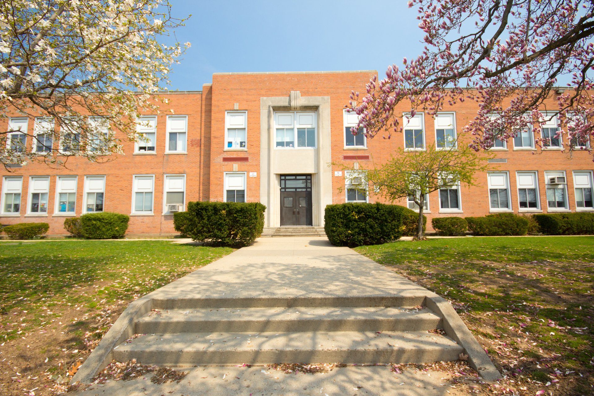 Brick school building with central entrance, steps, and trees in front under a clear blue sky