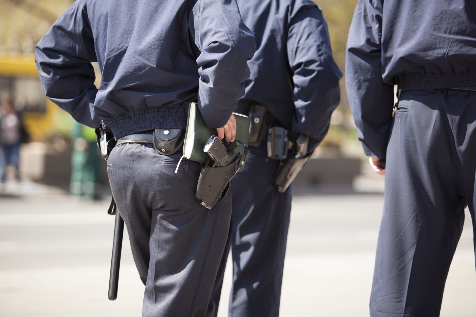 Three police officers in dark uniforms stand together outdoors, viewed from behind.
