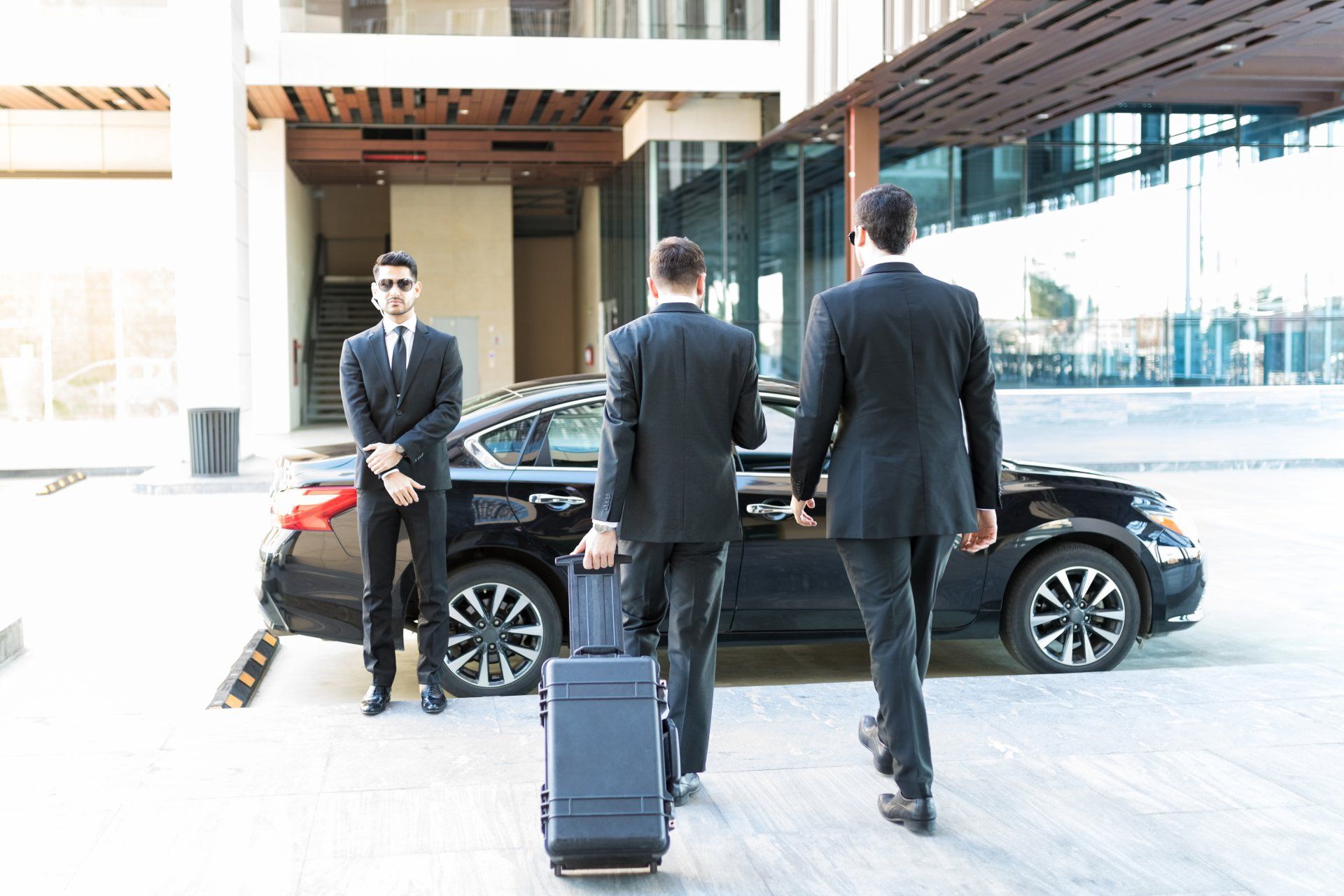 Three suited men with a suitcase near a black sedan at a modern building entrance